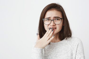 Portrait of young cute female in glasses, squinting and covering her mouth with hand, feeling pain