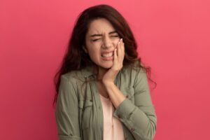 Aching young beautiful girl wearing olive green t-shirt putting hand on aching tooth isolated on pink wall