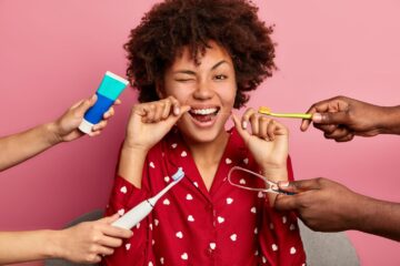 Happy curly young woman brushes teeth with tooth floss, cares about oral hygiene, surrounded with toothpaste, electric toothbrush and tongue cleaner