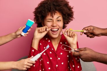Happy curly young woman brushes teeth with tooth floss, cares about oral hygiene, surrounded with toothpaste, electric toothbrush and tongue cleaner