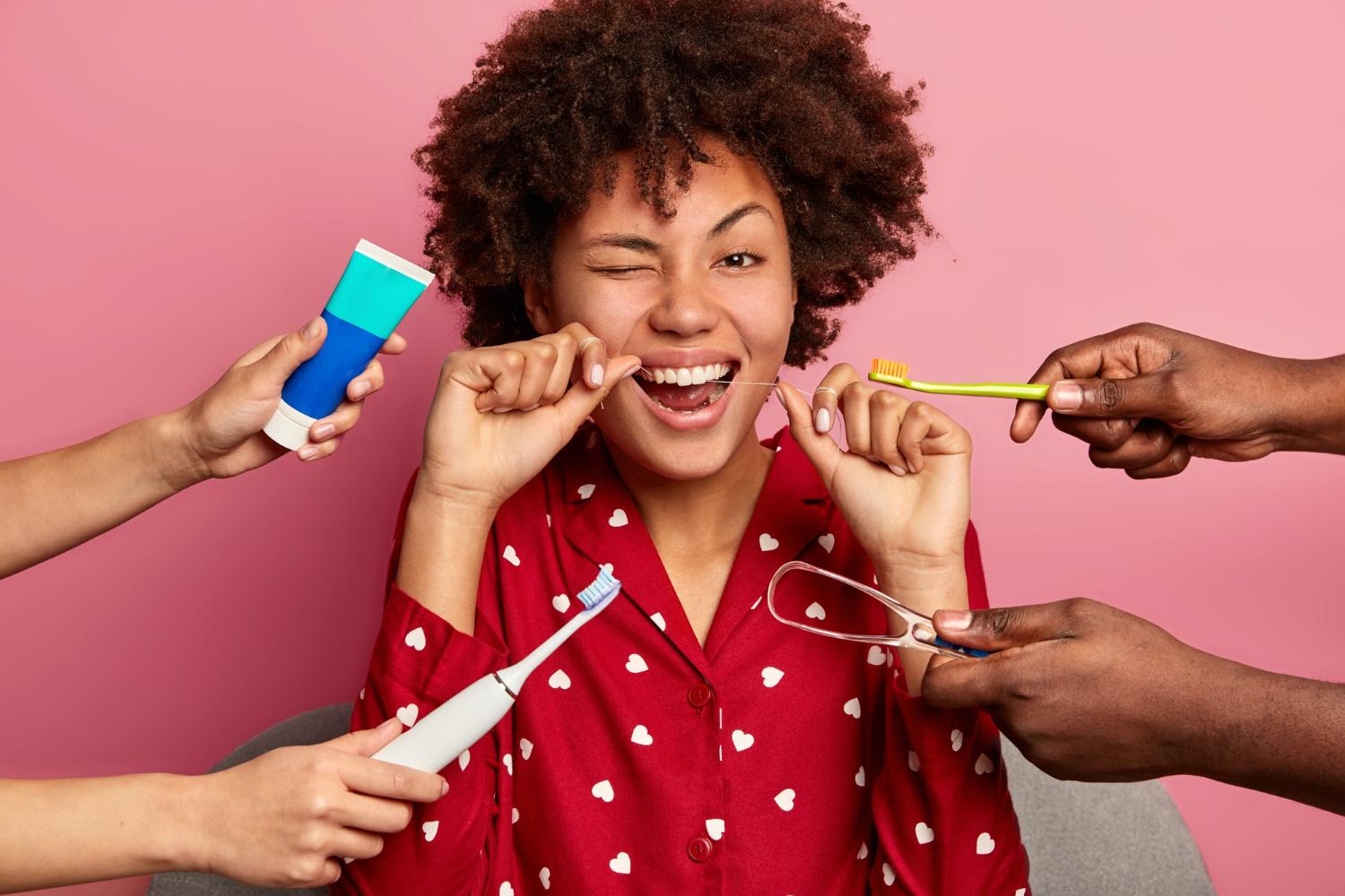Happy curly young woman brushes teeth with tooth floss, cares about oral hygiene, surrounded with toothpaste, electric toothbrush and tongue cleaner