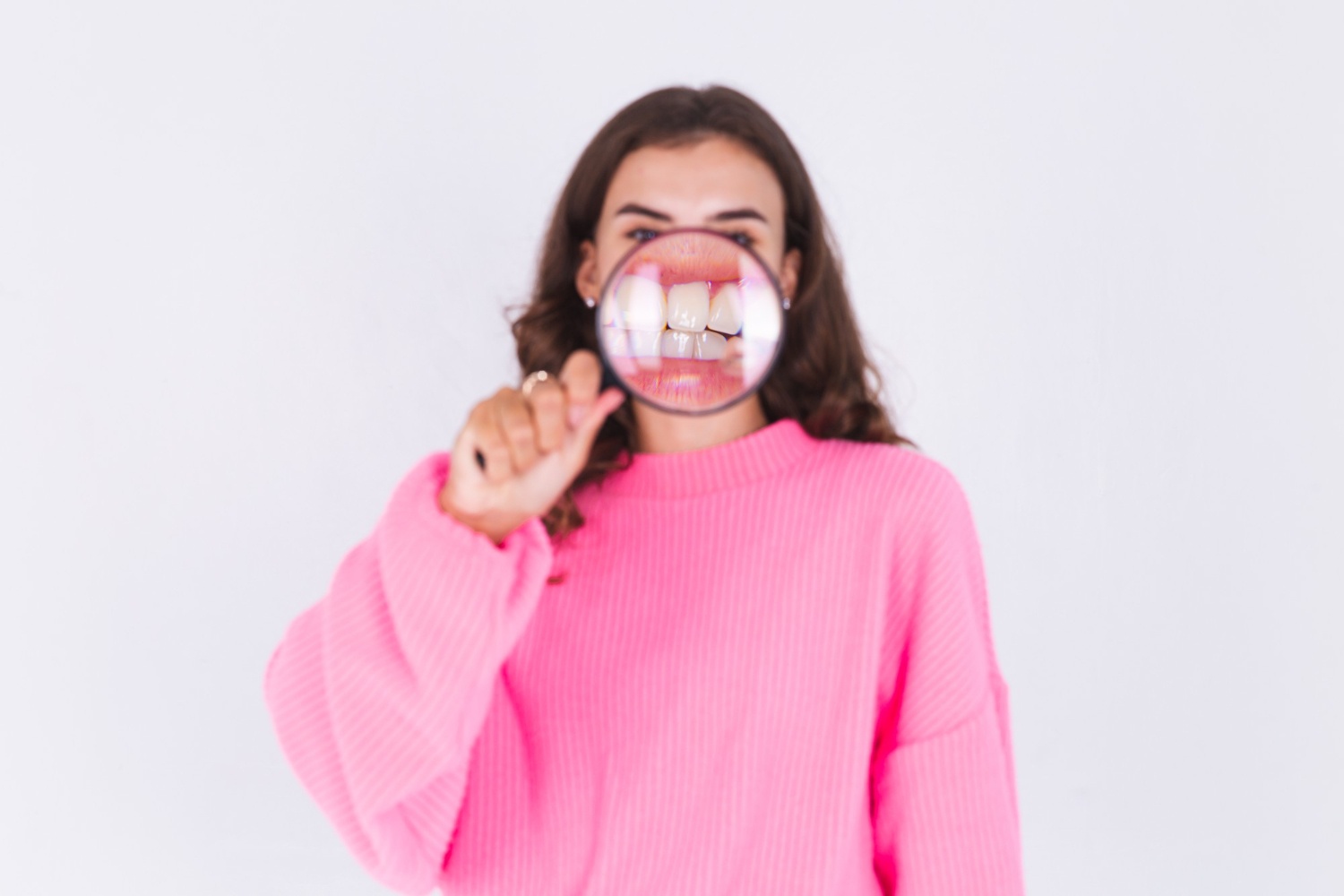 Young beautiful woman with freckles light makeup in sweater on white wall with magnifier shows white teeth perfect smile