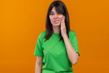 Annoyed young caucasian girl in green shirt holds face and looks at camera on isolated orange background