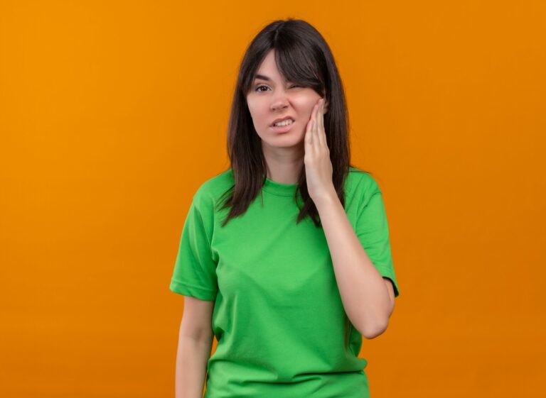 Annoyed young caucasian girl in green shirt holds face and looks at camera on isolated orange background