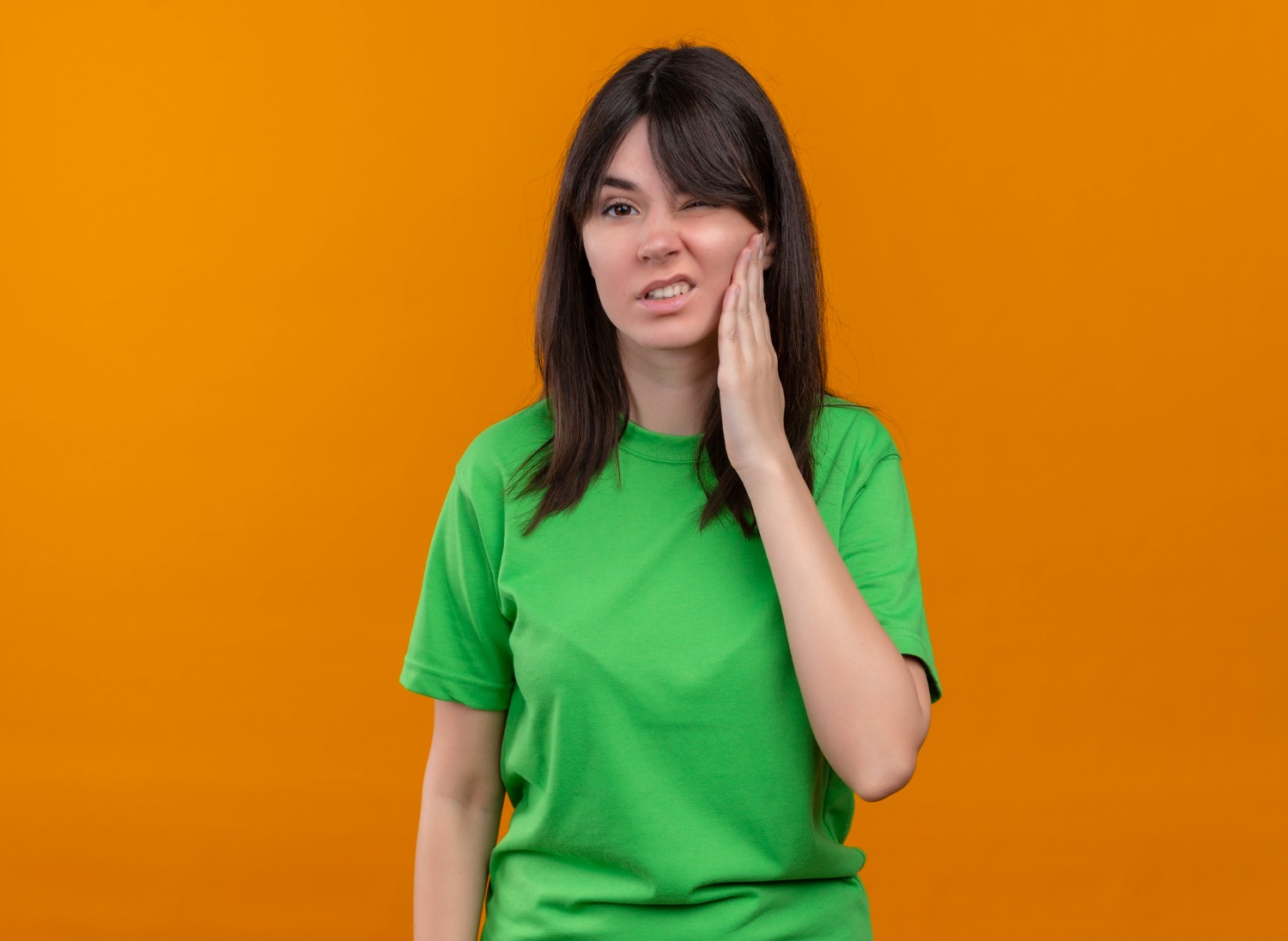 Annoyed young caucasian girl in green shirt holds face and looks at camera on isolated orange background
