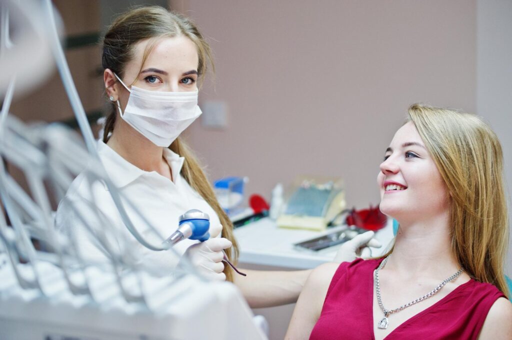 Attractive patient in redviolet dress laying on the dental chair while female dentist treating her teeth with special instruments